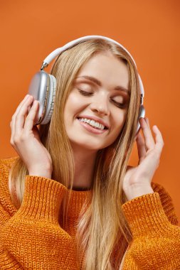 Parlak, sıcak süveter ve kulaklık giyen neşeli bir kadın turuncu gözleri kapalı müzik dinliyor.