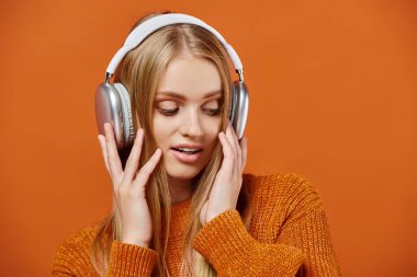Renkli örgü süveter ve kulaklıklarla turuncu arka planda müzik dinleyen çekici sarışın kadın.