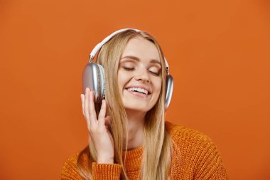 Parlak kazaklı ve kulaklıklı neşeli kadın turuncu arka planda kapalı gözlerle müzik dinliyor.