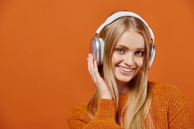 Turuncu kazaklı, kulaklıklı, müzik dinleyen ve kameraya bakan neşeli sarışın kadın.