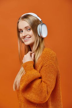 Turuncu kışlık süveter ve kulaklık giyen neşeli sarışın kadın müzik dinliyor ve kameraya bakıyor.
