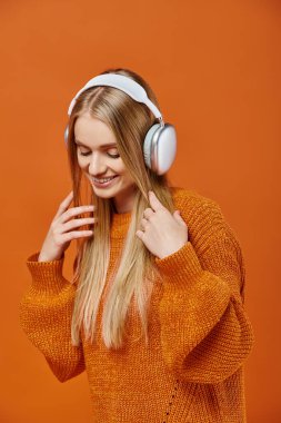 Sıcak turuncu süveter ve kulaklık takan gülümseyen sarışın kadın parlak arka planda müzik dinliyor.