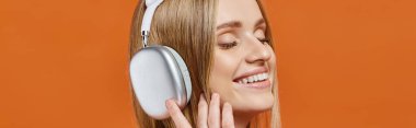 Mutlu sarışın kadın portresi. Kulaklıkla müzik dinliyor. Gözleri turuncu, afişi kapalı.