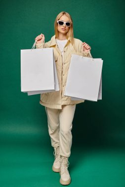 fashionable blonde woman in winter attire and sunglasses showing hopping bags on green backdrop clipart