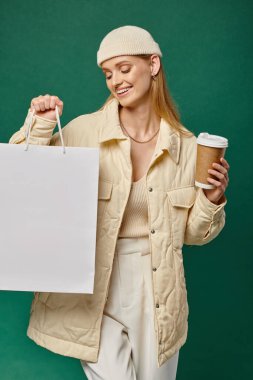 happy woman in warm winter clothes with paper cup looking at shopping bag on green, seasonal sales clipart