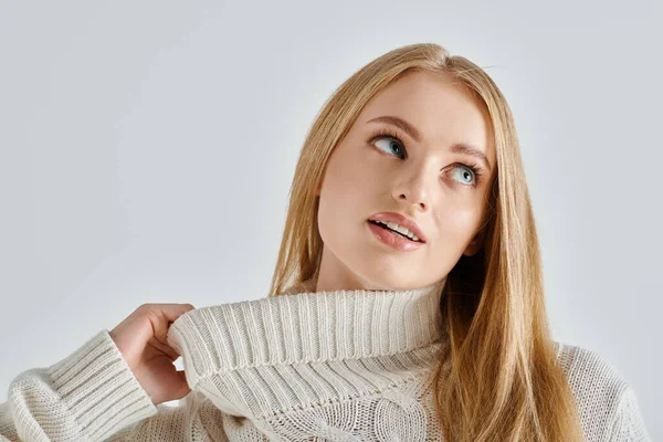 Stock image charming and dreamy woman stretching collar of white knitted sweater and looking away on grey