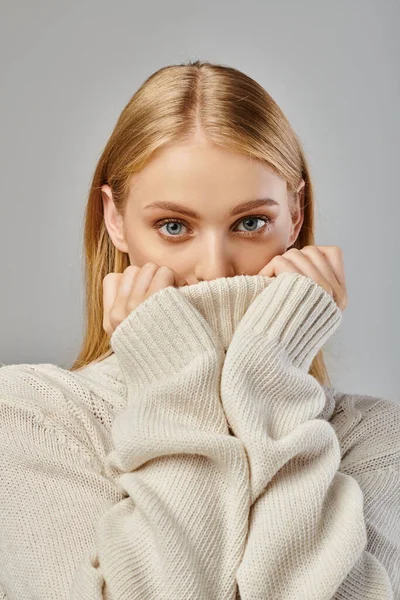 stock image young blonde woman obscuring face with warm winter sweater on grey backdrop, sophisticated charm