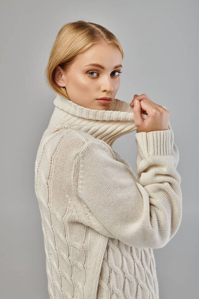 young blonde woman in warm knitted sweater with high collar looking at camera on grey, winter trend