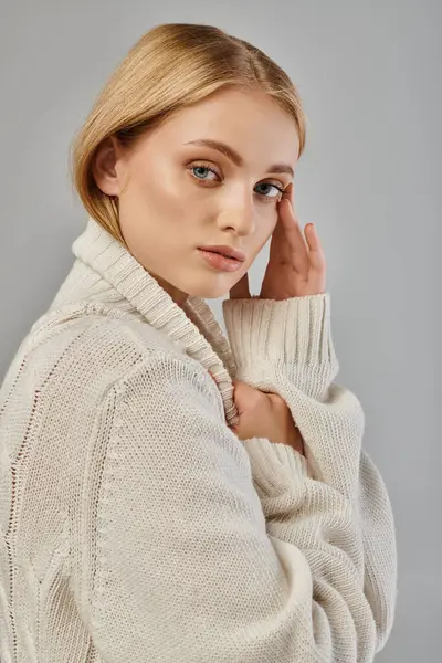 Beyaz örgü süveter giyen çekici sarışın kadın yüzünün yanında el ele tutuşuyor ve gri kameraya bakıyor.