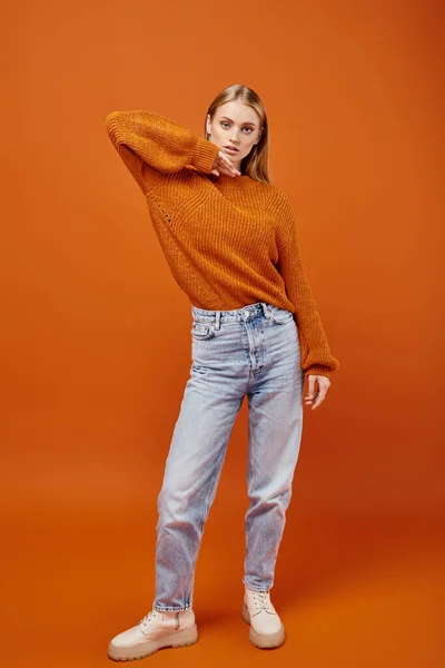 Stock image trendy blonde woman in warm knitted sweater and blue jeans on orange backdrop, winter fashion