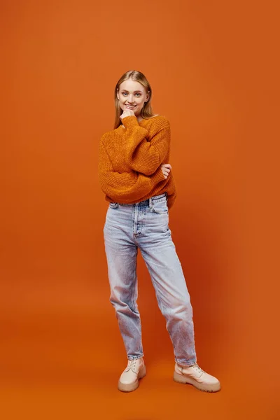 stock image joyful blonde woman in orange winter sweater and blue jeans smiling at camera on bright backdrop