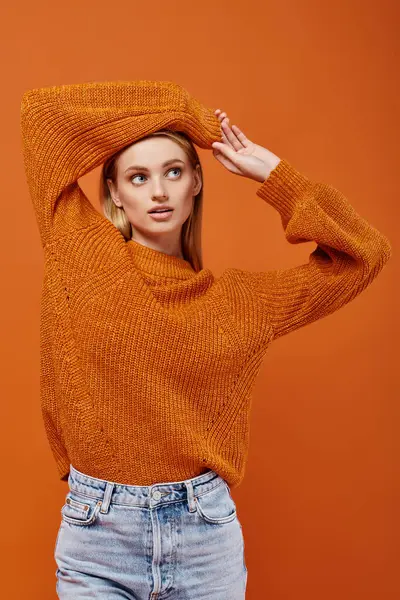 stock image young blonde woman in orange knitted sweater posing with arms above head on orange, winter vibes