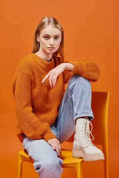 stock image blonde woman in warm stylish sweater and blue jeans sitting on chair and looking at camera on orange