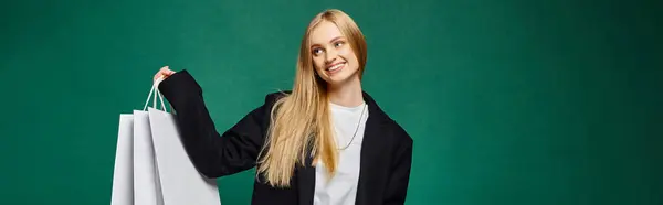 stock image happy blonde woman in black trendy coat posing with white shopping bags on green, horizontal banner