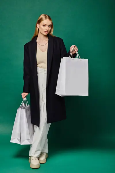 stock image full length of stylish blonde woman in black coat posing with white shopping  bags on green