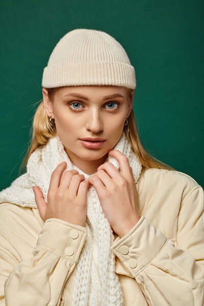 young woman in warm jacket and beanie hat with white scarf looking at camera on green, winter trend