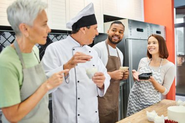 cheerful multicultural people in aprons talking to mature chef in white hat, cooking courses clipart