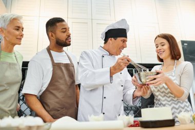neşeli olgun aşçı ders sırasında hamur çırpıyor ve çeşitli öğrencilere gülümsüyor, yemek kursları veriyor.