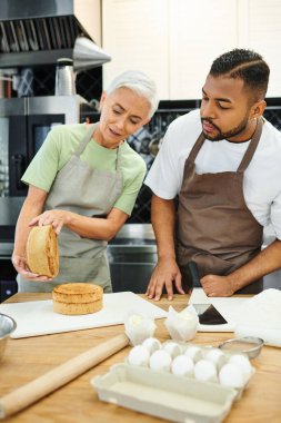 mature beautiful woman and african american man with aprons baking delicious cake, cooking courses clipart