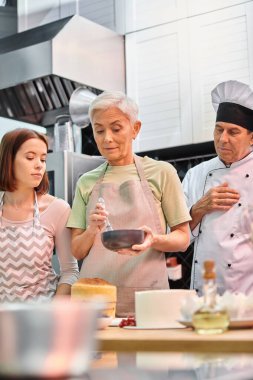 mature woman in apron learning how to use silicone brush on cake next to her young friend and chef clipart