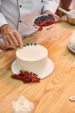 cropped view of mature chef showing his student how to decorate cake with berries, cooking courses clipart