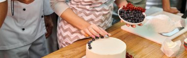 cropped view of young woman decorating cake with berries next to her friend and mature chef, banner clipart