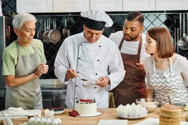 Beyaz şapkalı neşeli olgun aşçı ders sırasında çeşitli öğrencilerinin yanında pastasını süslüyor.