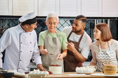 mature jolly woman decorating cake next to her multicultural friends and chef, cooking courses clipart