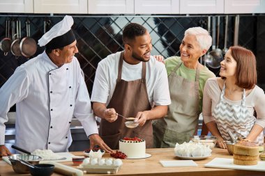 Önlüklü neşeli Afrikalı Amerikalı adam pasta süslüyor ve arkadaşlarına ve aşçısına gülümsüyor.