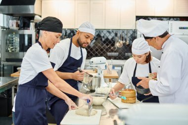 Önlüklü ve şapkalı iyi görünümlü şefler aşçıbaşıyla birlikte pasta ve şekerleme üzerinde çalışıyorlar.