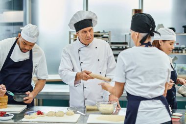 Beyaz şapkalı yakışıklı aşçı kadın aşçısına merdane veriyor.