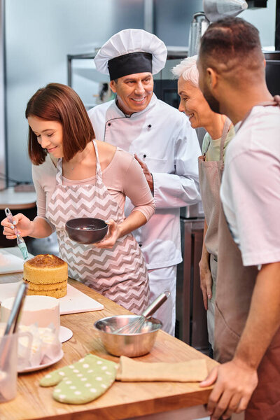 young cheerful woman brushing cake with syrup while her diverse friends and chef talking actively