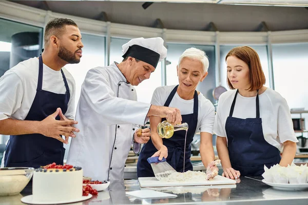 cheerful mature chef in white hat pouring some oil in dough next to his diverse colleagues