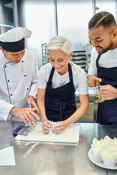 stock image cheerful mature female chef working with dough next to african american colleague and chief cook