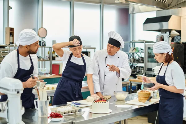 mature chief cook in white hat explaining how to bake to his multicultural hard working chefs