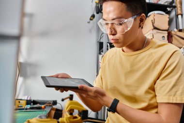 Gözlüklü dikkatli Asyalı tamirci atölyede kırık dokunmatik ekranlı dijital tablet tutuyor.