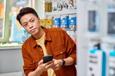 Asyalı girişimci cep telefonu tutuyor ve kendi elektronik mağazasına bakıyor, küçük işletme.