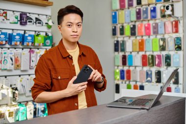 Cep telefonlu kendine güvenen Asyalı satıcı özel dükkandaki bilgisayarın yanındaki kameraya bakıyor.