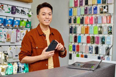 Akıllı telefonlu neşeli Asyalı adam elektronik mağazasında bilgisayarın yanındaki kameraya bakıyor.