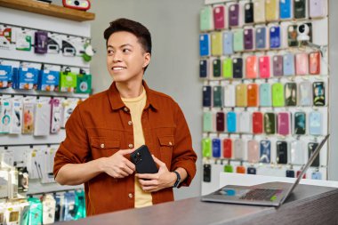Cep telefonlu mutlu Asyalı işadamı özel elektronik mağazasında dizüstü bilgisayarın yanından uzağa bakıyor.