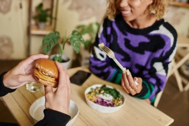 cropped view of young and diverse couple enjoying vegan meal in cafe, burger with tofu and salad clipart