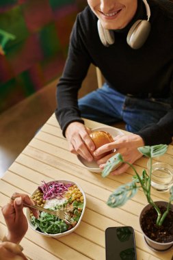 overhead view of young diverse couple enjoying vegan meal in cafe, burger with tofu and salad bowl clipart