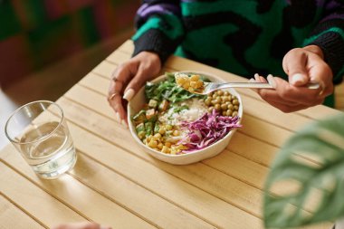 Afro-Amerikan kadının kesilmiş fotoğrafı. Elinde mısır çatalıyla kafede vegan kasesi yerken çekilmiş.