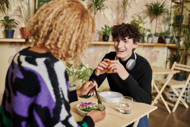 Afro-Amerikan kız arkadaşının yanında tofu burger yiyen mutlu kıvırcık adam vejetaryen kafede salata yiyor.
