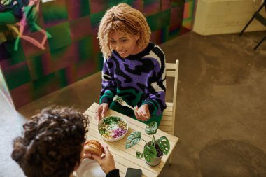 Tel takmış neşeli Afro-Amerikalı kadın kafede vejetaryen burgerli bir adamın yanında lezzetli salata yiyor.