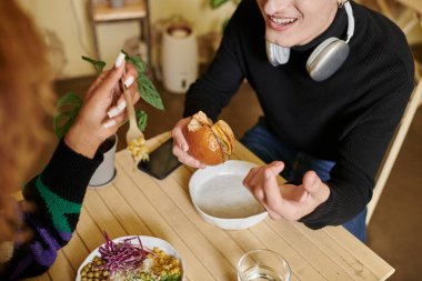 cropped shot of happy man in wireless headphones eating vegan tofu burger near woman in cafe clipart