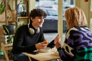 Kablosuz kulaklıklı mutlu kıvırcık adam elinde tofu burger ve vejetaryen kafede kızla sohbet ediyor.
