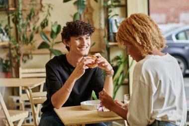 Afro-Amerikan kızının yanında tofu burger tutan memnun ve kıvırcık adam vejetaryen kafede salata yiyor.