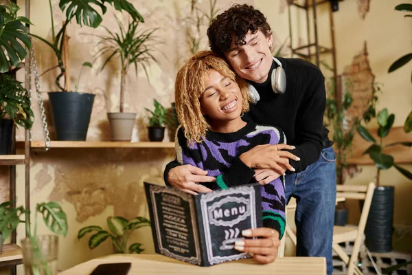 stock image affectionate and curly young man embracing african american girlfriend near menu in vegan cafe