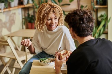 Kafede vejetaryen burger yiyen kıvırcık erkek arkadaşına bakan, diş teli takan mutlu Afro-Amerikalı kız.